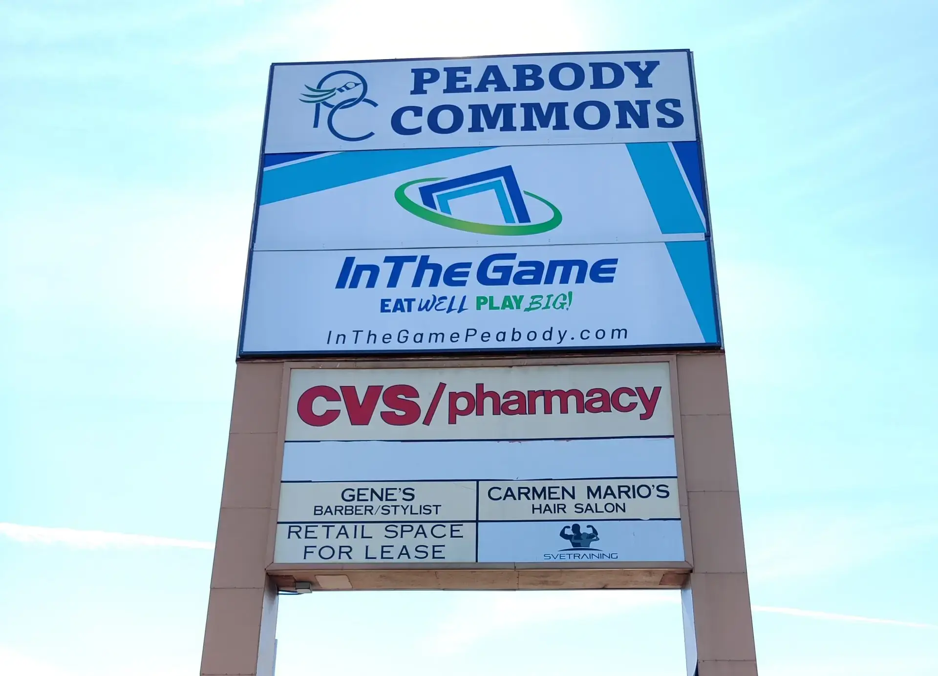 Signage for Peabody Commons featuring InTheGame, CVS/pharmacy, Gene's Barber/Stylist, and Carmen Mario's Hair Salon, against a clear blue sky.