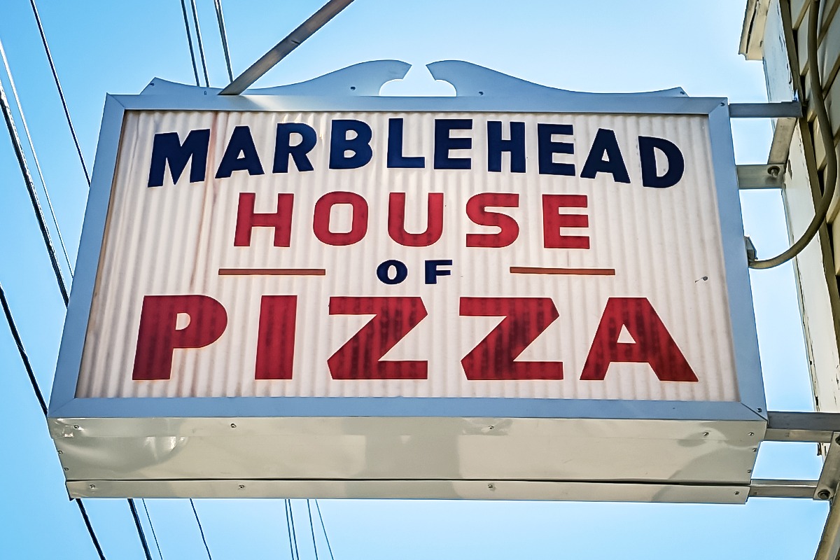 Illuminated sign for Marblehead House of Pizza featuring bold red and blue lettering, showcasing the business name against a bright sky background, emphasizing visibility and attraction for customers.