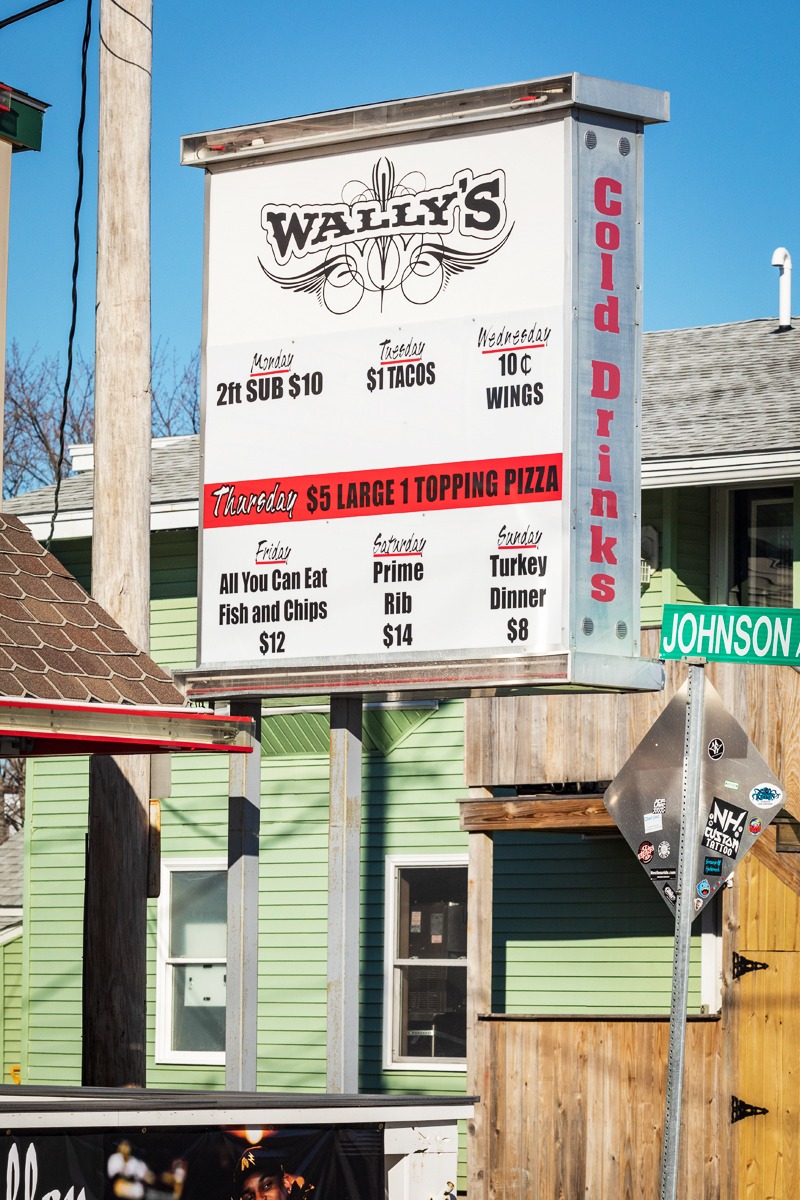 Wally's illuminated menu board displaying daily specials including $5 large one topping pizza on Thursdays, located in a vibrant New England setting.