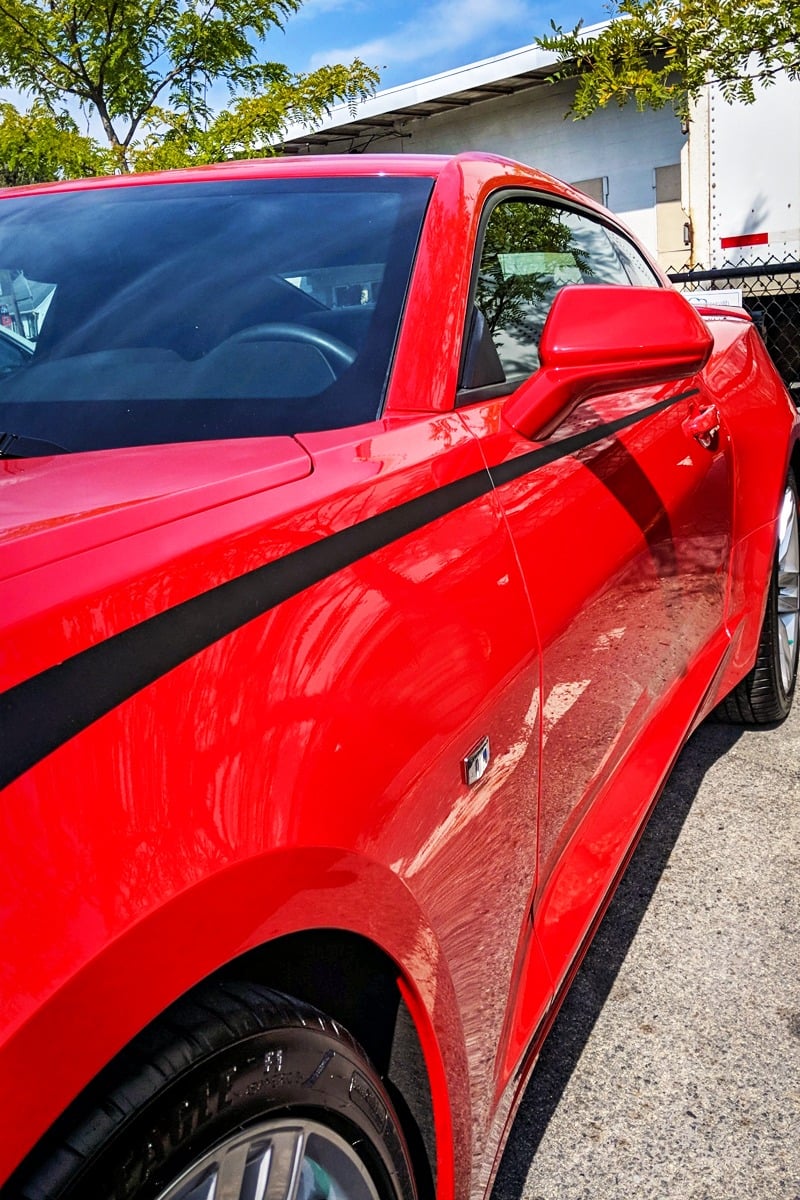Red Camaro with black racing stripe, showcasing vehicle graphics for branding, parked in a well-lit outdoor setting.