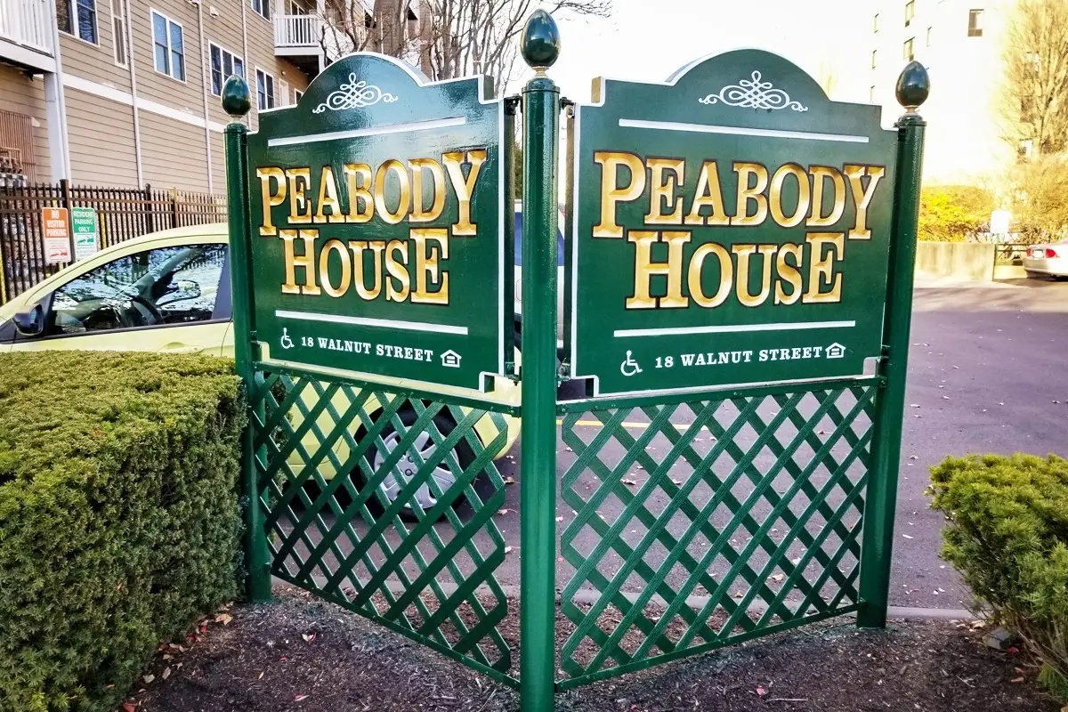 Monument sign for Peabody House, featuring green design with gold lettering, displaying the address "18 Walnut Street" and accessibility symbol, surrounded by landscaping.