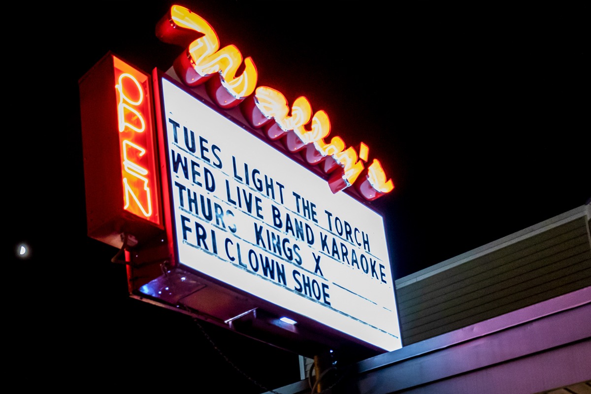 Wally's neon sign displaying events schedule with "Open" indicator, showcasing vibrant colors and illuminated lettering against a nighttime backdrop.