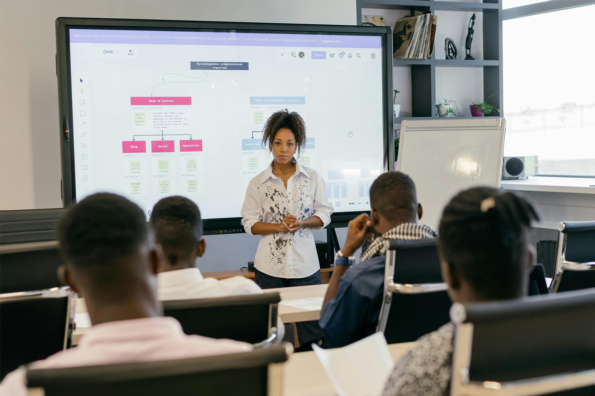 Digital presentation in a meeting room with a speaker addressing an audience, featuring a large screen displaying a flowchart and notes.