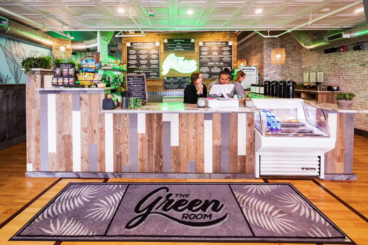 Counter area of 'The Green Room' café featuring two staff members, a menu board, and a decorative rug, emphasizing a welcoming atmosphere for customers seeking smoothies and snacks.