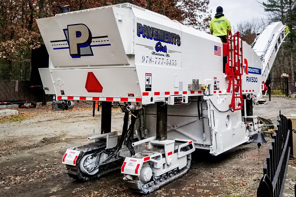 Heavy-duty pavement milling machine with tracks, featuring branding and contact information, positioned on a gravel surface in a construction setting.
