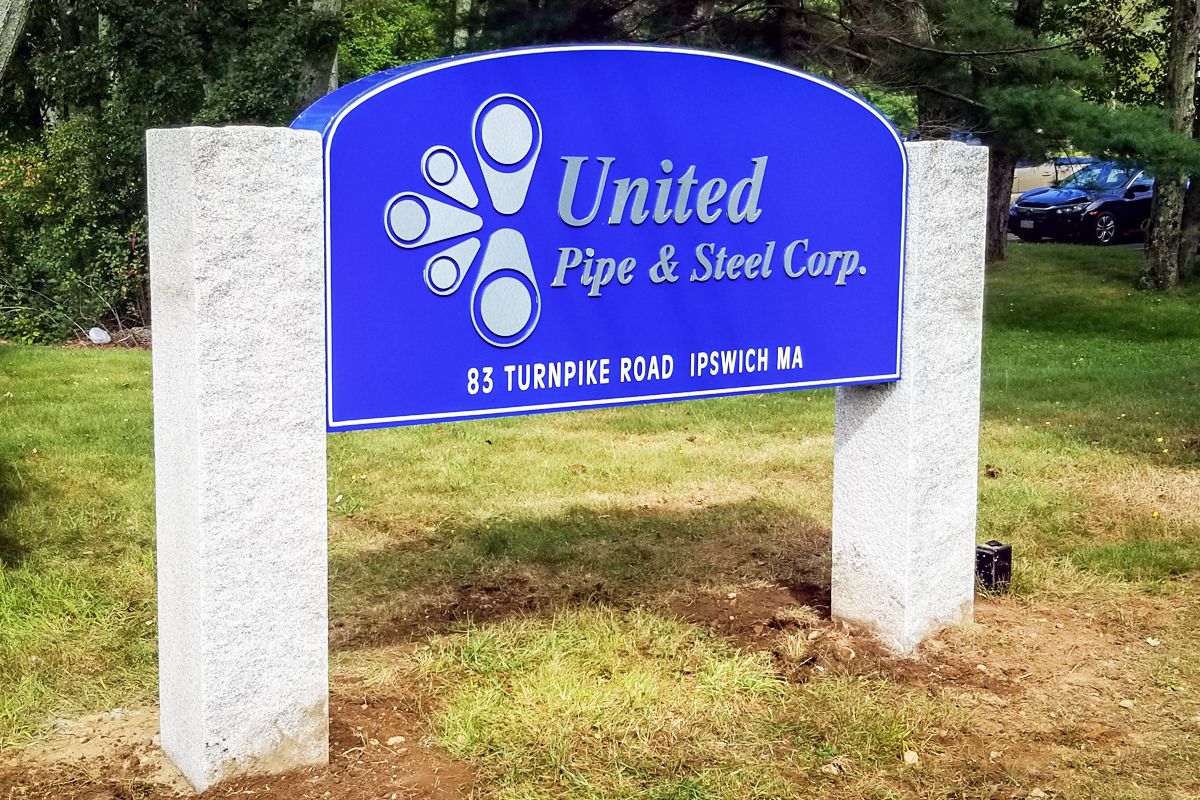 Monument sign for United Pipe & Steel Corp, featuring blue background, company name, and address at 83 Turnpike Road, Ipswich MA, surrounded by greenery.