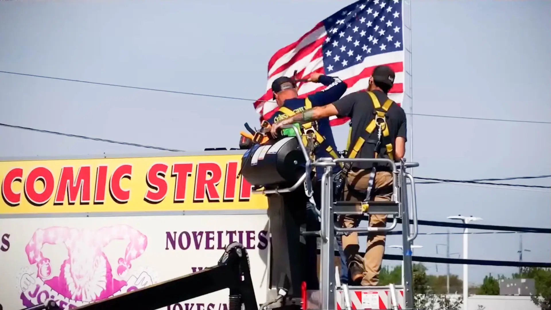 Team of workers on a lift installing a tall sign for a business, featuring an American flag and the words "Comic Strip" prominently displayed.
