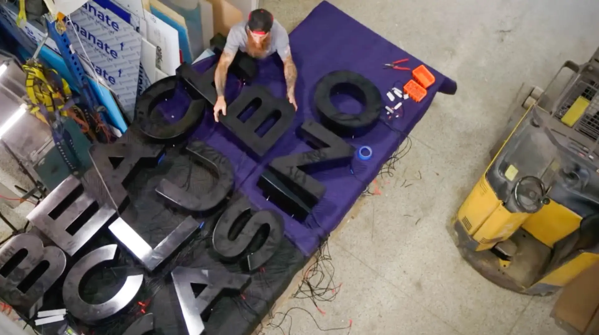 Top-down view of a worker arranging large, black sign letters on a purple table, with tools and materials for sign-making visible, in a workshop setting.