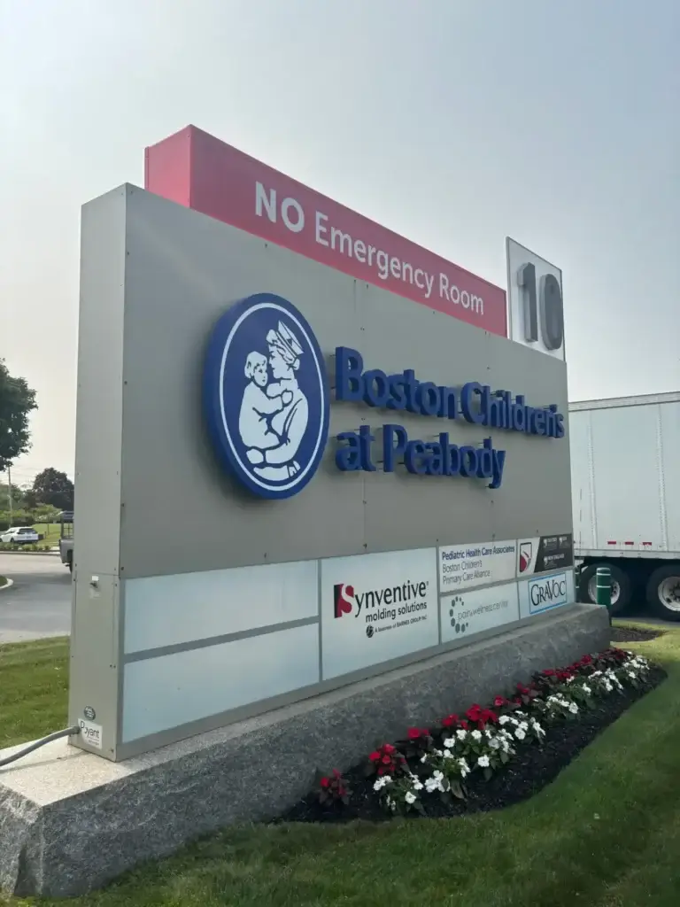 Sign for Boston Children's at Peabody, featuring logo of a mother and child, with "NO Emergency Room" text prominently displayed above, surrounded by flower beds and nearby vehicles.