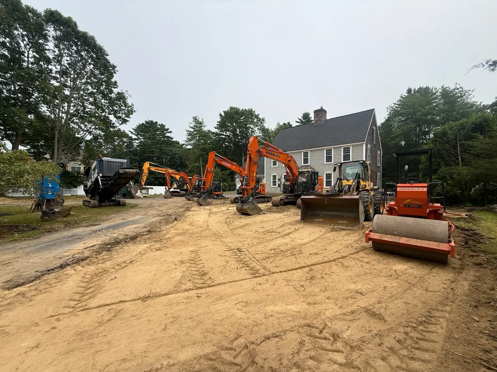Construction site featuring heavy machinery including excavators, a bulldozer, and a roller on sandy terrain, adjacent to a house, illustrating active construction zone signage and equipment for project compliance.