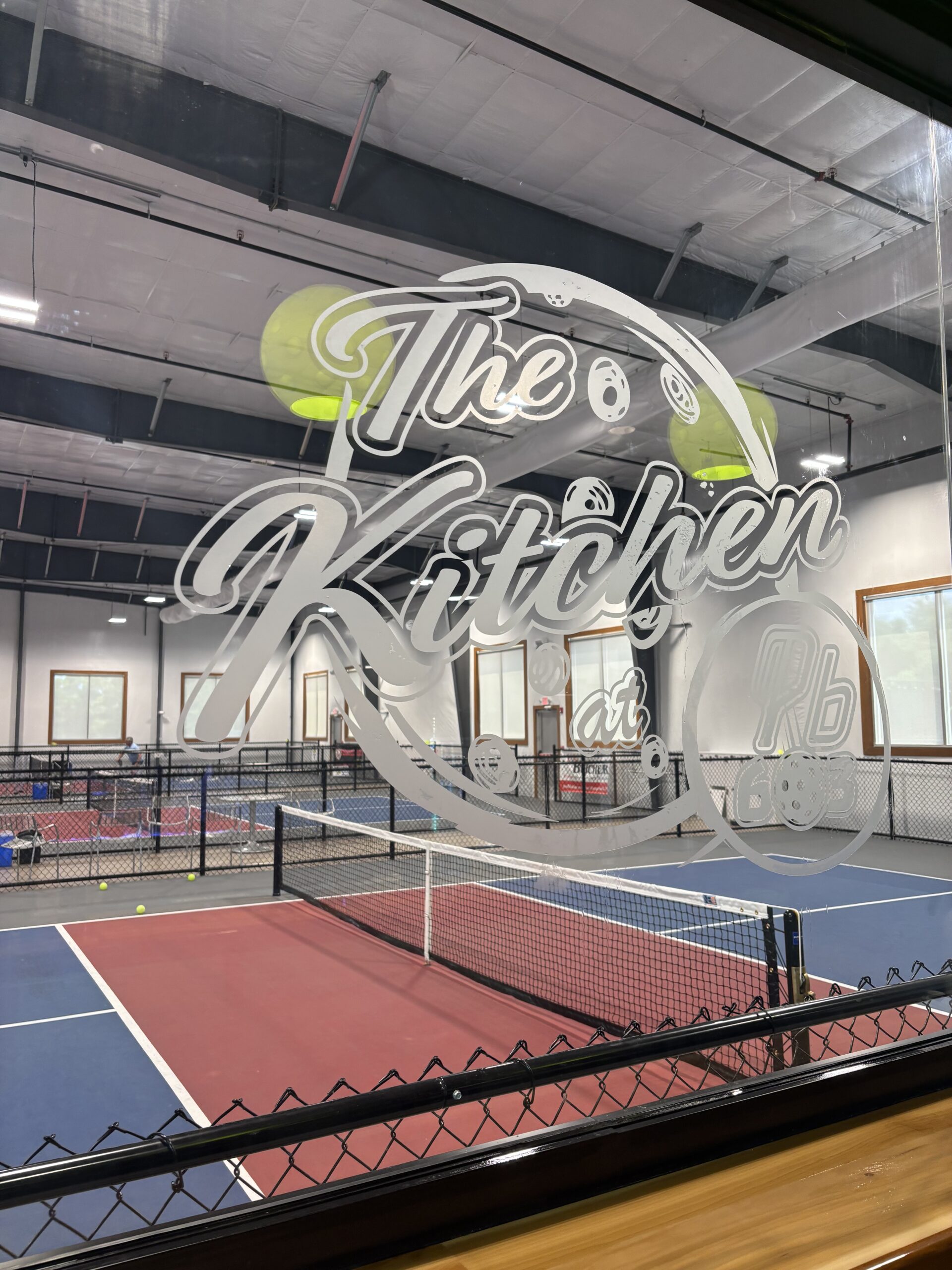 Vinyl lettering on glass window displaying "The Kitchen at" in a sports facility, with tennis courts and equipment visible in the background.
