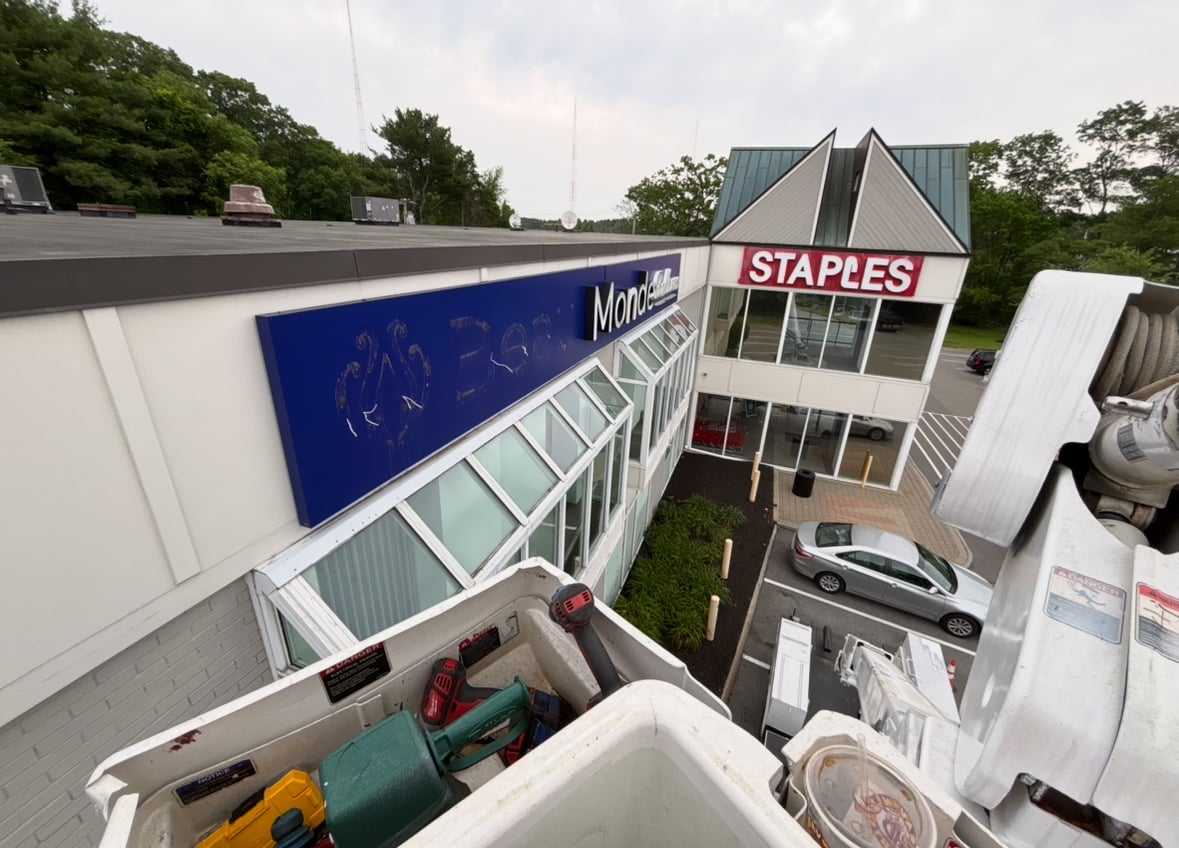 Corporate logo sign on building facade featuring blue background and stylized text, with adjacent Staples store visible, showcasing professional branding and signage solutions.