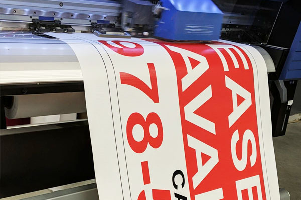 Large vinyl banner printing process featuring bold red text reading "EASE AVAILABLE" and a contact number, showcasing high-resolution digital printing technology used by Signs East for customized signage solutions.