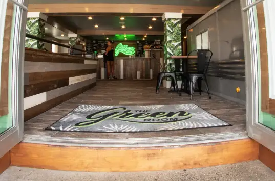 Entrance mat featuring "The Green Room" logo, surrounded by a welcoming indoor space with wooden flooring and greenery, highlighting branded floor graphics for retail environments.