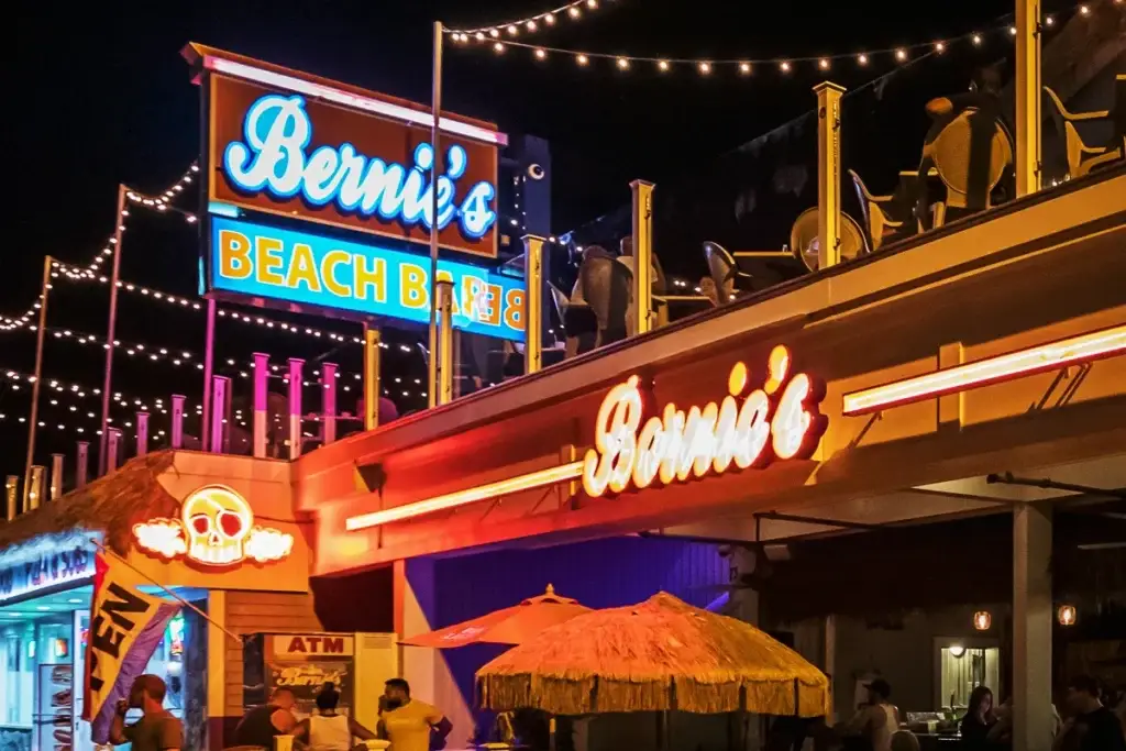 Bernie's Beach Bar exterior at night with vibrant neon signage, outdoor seating, and festive string lights, illustrating a lively beachside atmosphere.