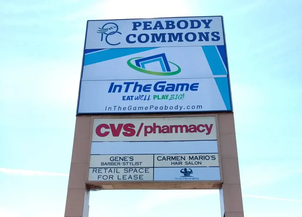 Peabody Commons shopping center sign featuring In The Game, CVS/pharmacy, Gene's Barber Stylist, and Carmen Mario's Hair Salon, highlighting local business options.