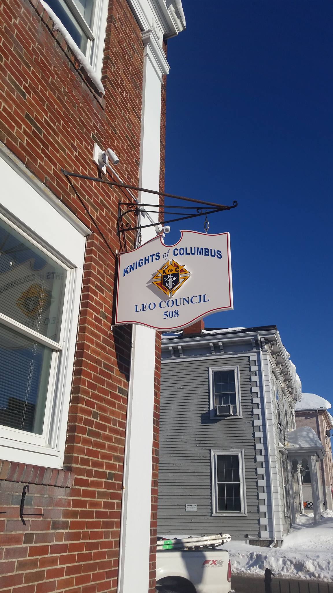 Knights of Columbus Leo Council 508 sign hanging from a brick building, showcasing a colorful emblem and clear text, against a bright blue sky.