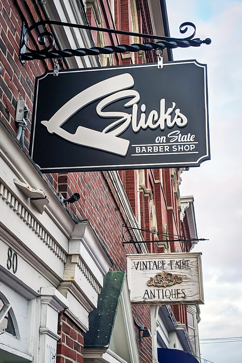 Sidewalk frame sign advertising "Slick's on State Barber Shop" and "Vintage Faire Antiques," showcasing double-sided panels for outdoor visibility, set against a brick storefront.