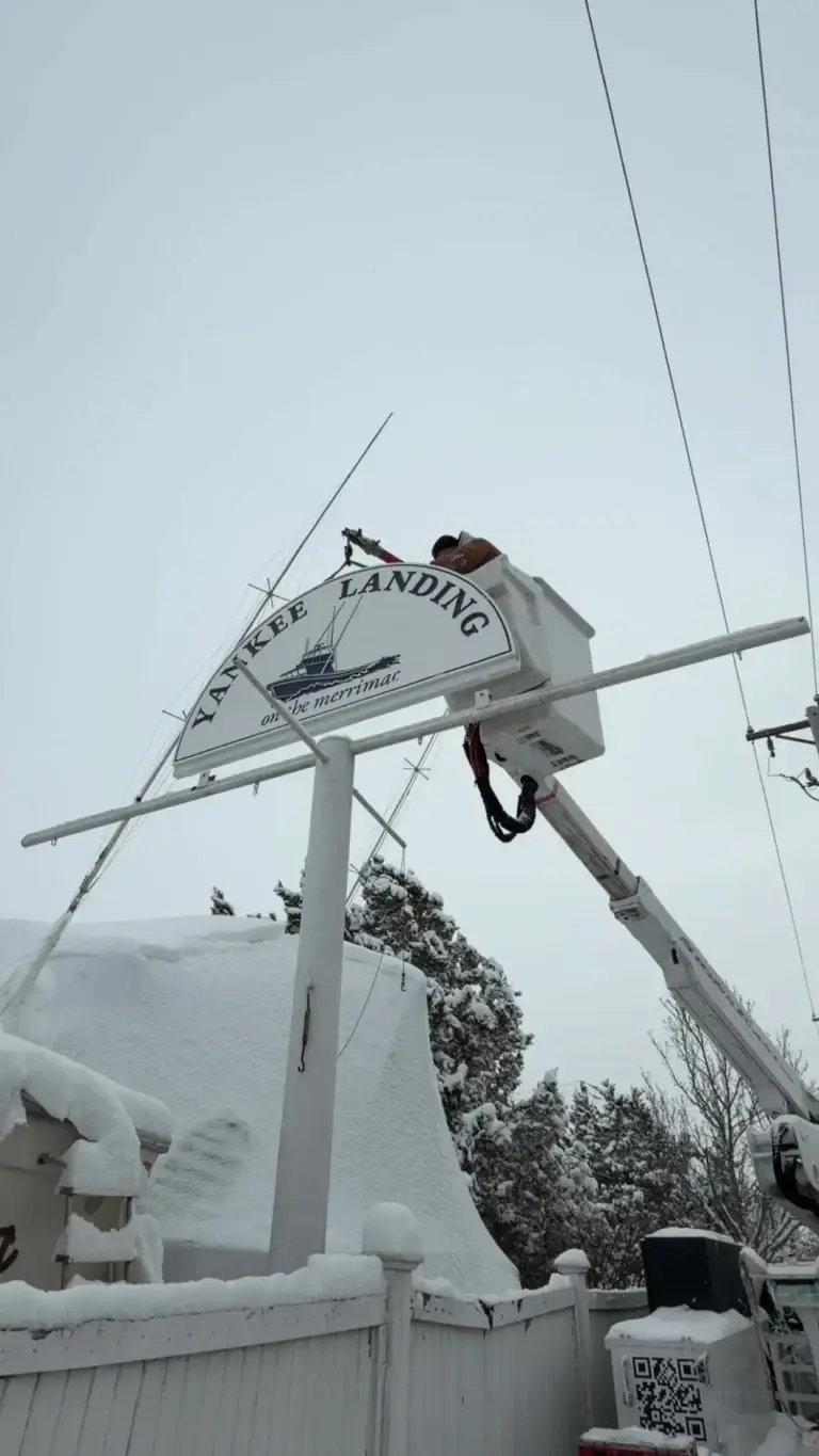 Worker on cherry picker installing a sign at Yankee Landing amid snow.